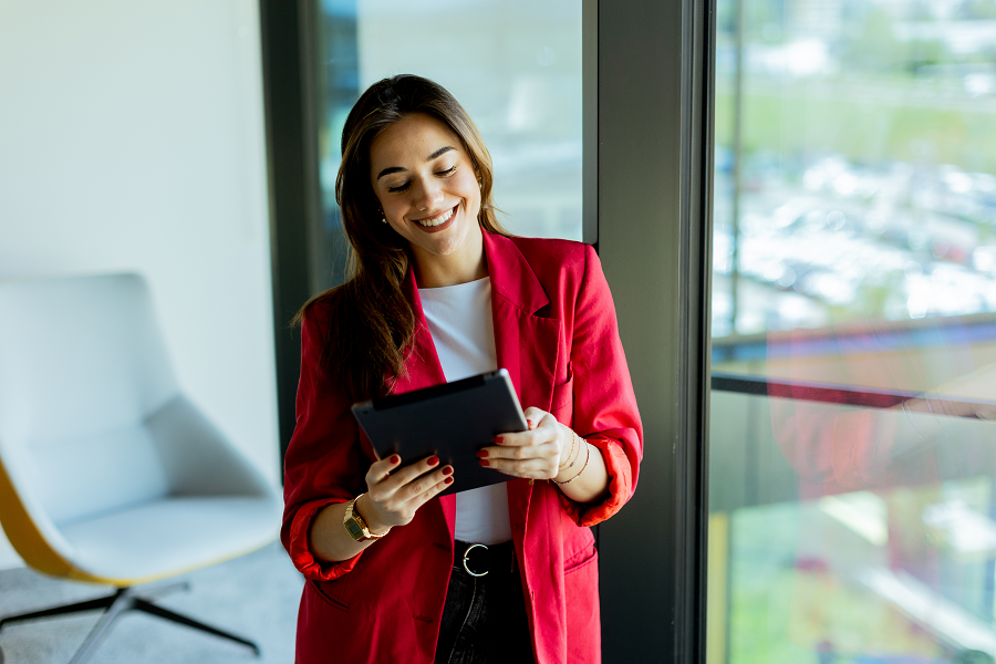 A woman in a red blazer, white top and jeans uses a tablet at work.